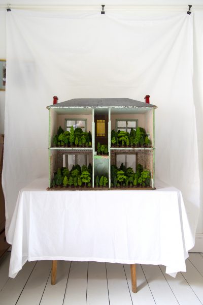 A dollshouse filled with model trees stands on a table with a white cloth